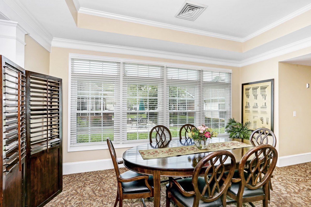 A bright dining room with a large table and several chairs, featuring ample natural light from the wide windows adorned with blinds.