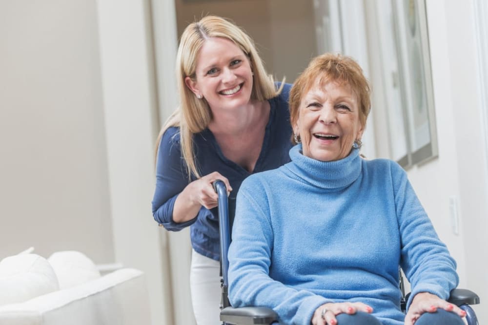 A caregiver happily assists an elderly woman in a wheelchair indoors.