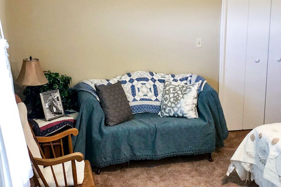 A cozy living room corner featuring a small sofa draped in blankets and pillows, with a chair nearby.