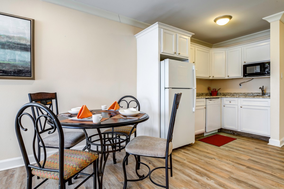 A cozy kitchen and dining area featuring a round table set for four, with a refrigerator and dishwasher in the background.