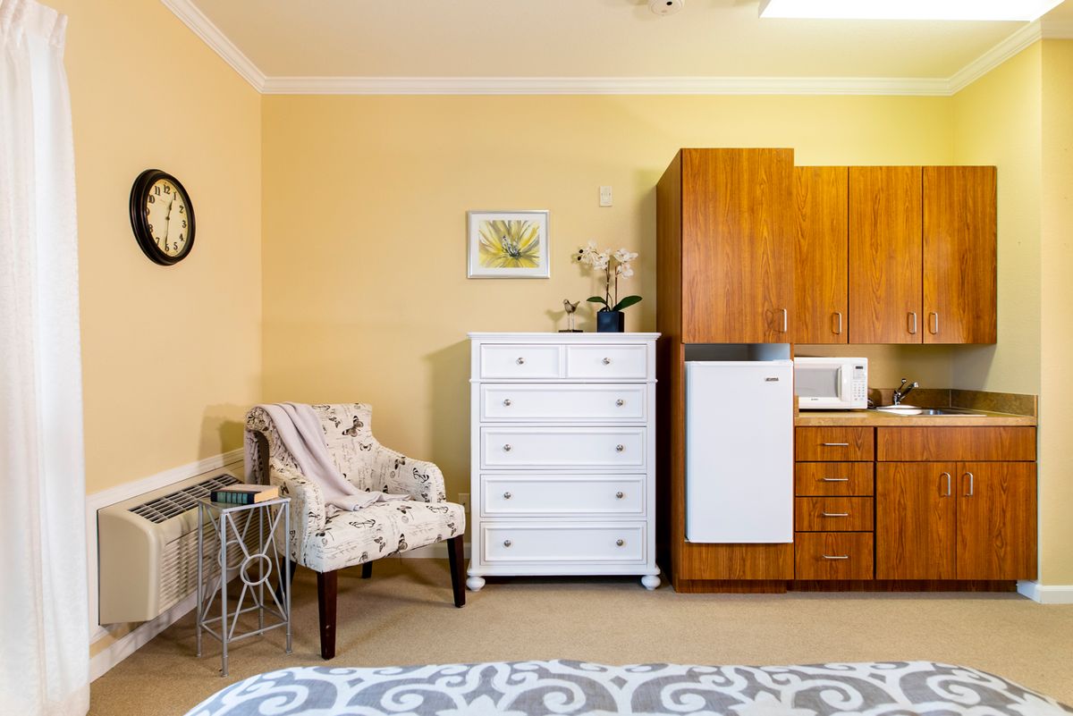 A cozy room interior featuring a white dresser, patterned chair, and kitchen corner with a mini fridge and microwave.