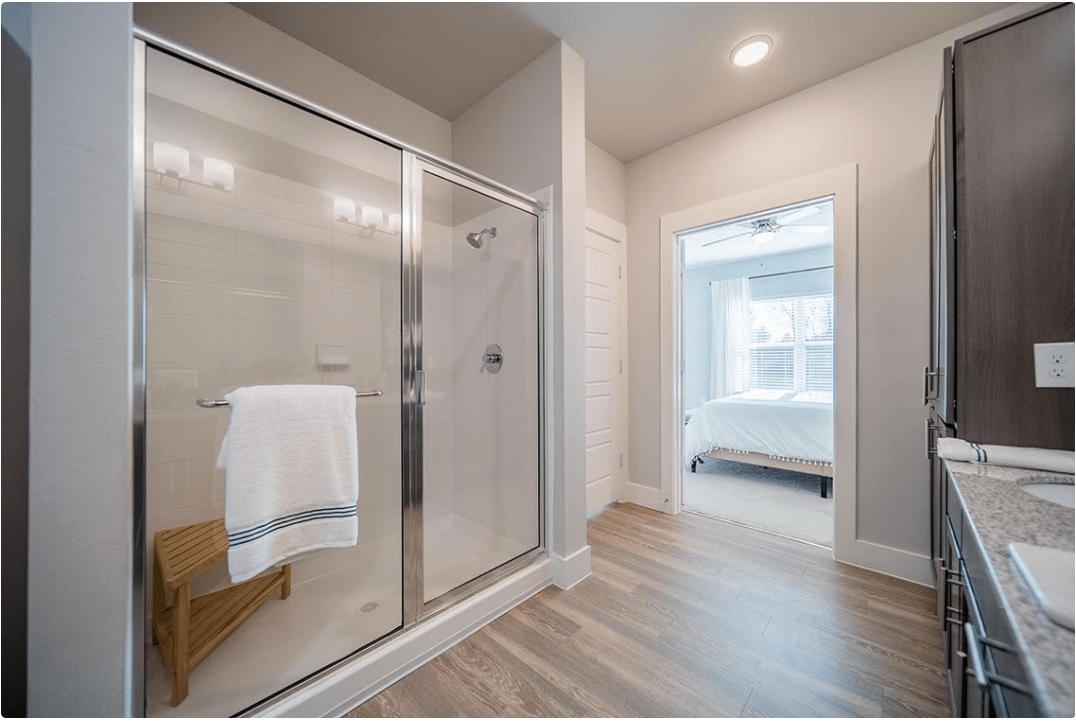 A modern bathroom with a glass shower and wood flooring leading to a bright bedroom.