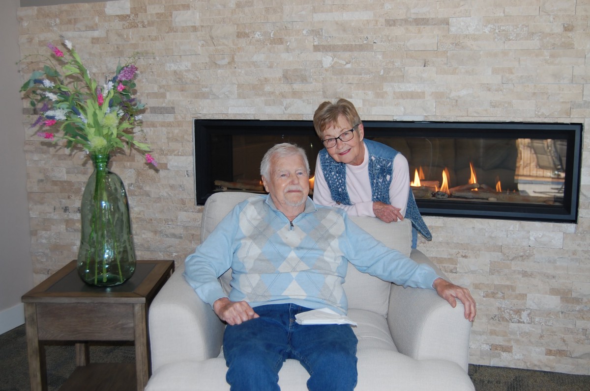An elderly man sitting in a comfortable chair with a woman standing beside him, indoors by a fireplace.