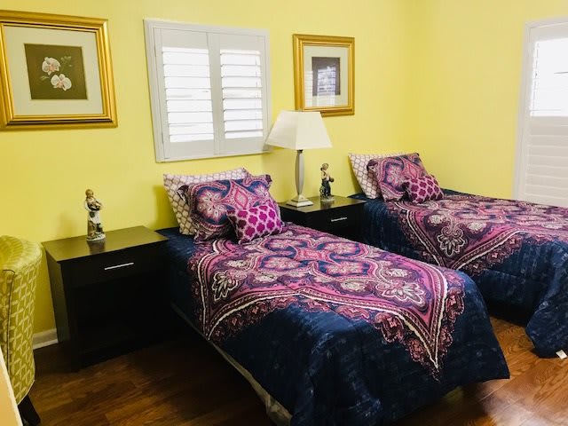 A cozy bedroom with twin beds adorned in colorful bedding, set against sunny yellow walls with artwork and a window featuring shutters.