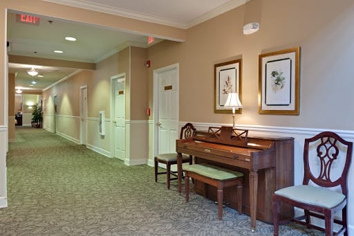 A cozy hallway features a wooden piano accompanied by chairs, framed artwork on the walls, and soft carpeting.
