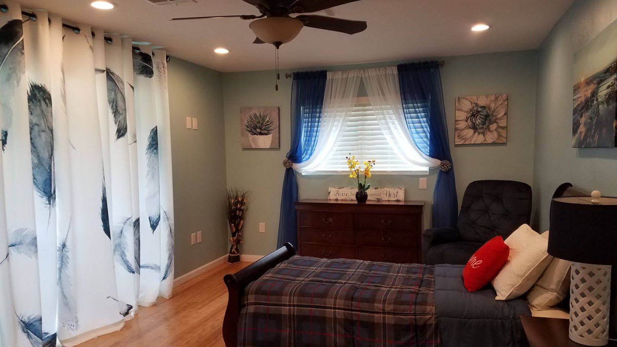 A cozy bedroom featuring a bed with plaid bedding, a window with blue curtains, and a dresser topped with flowers.