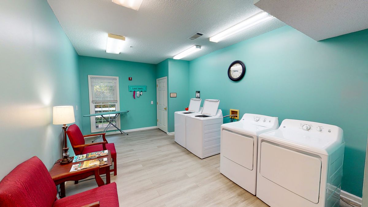 A brightly lit laundry room with turquoise walls, featuring washing machines and dryers, red chairs, and an ironing board.
