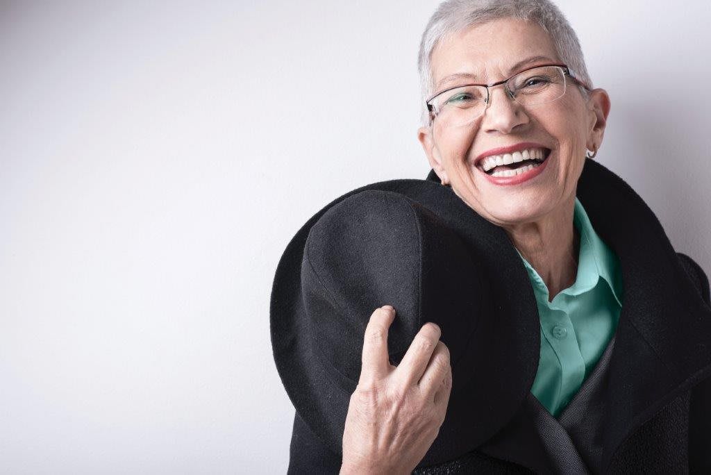 A cheerful elderly woman with glasses holding a hat and smiling widely.