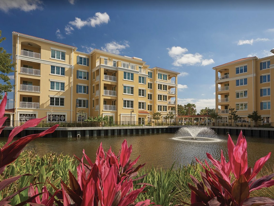 A scenic view of modern apartment buildings with a lake and fountain in the foreground, surrounded by vibrant plants.
