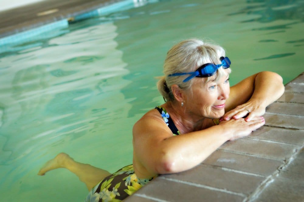 An elderly woman relaxing at the edge of a swimming pool, wearing goggles.
