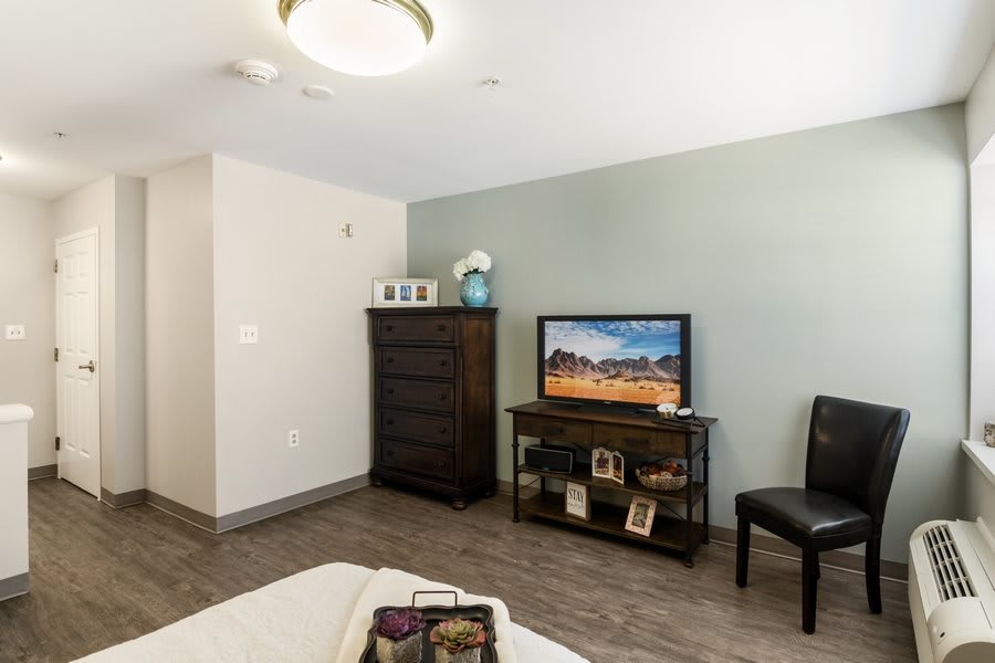 A minimalist bedroom interior featuring a television on a stand, dresser, and a chair.