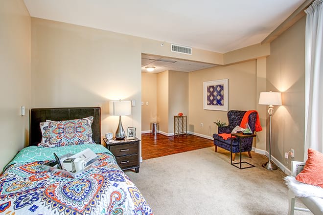 A cozy bedroom with colorful decor, featuring a bed with patterned bedding, a side table with a lamp, and a comfortable chair in the corner.
