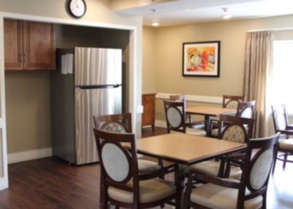 A cozy dining area with tables, chairs, and a refrigerator, featuring warm lighting and artwork on the wall.