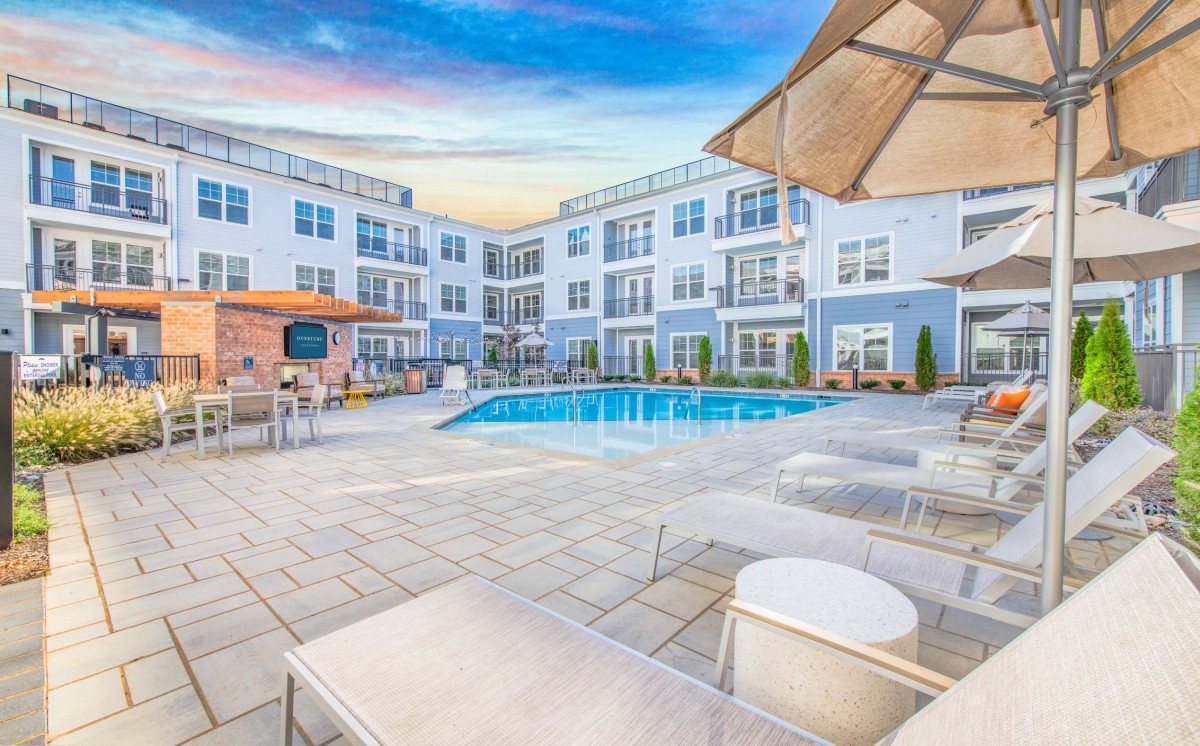 A modern apartment complex featuring a central swimming pool surrounded by outdoor lounge areas and patio furniture at sunset.