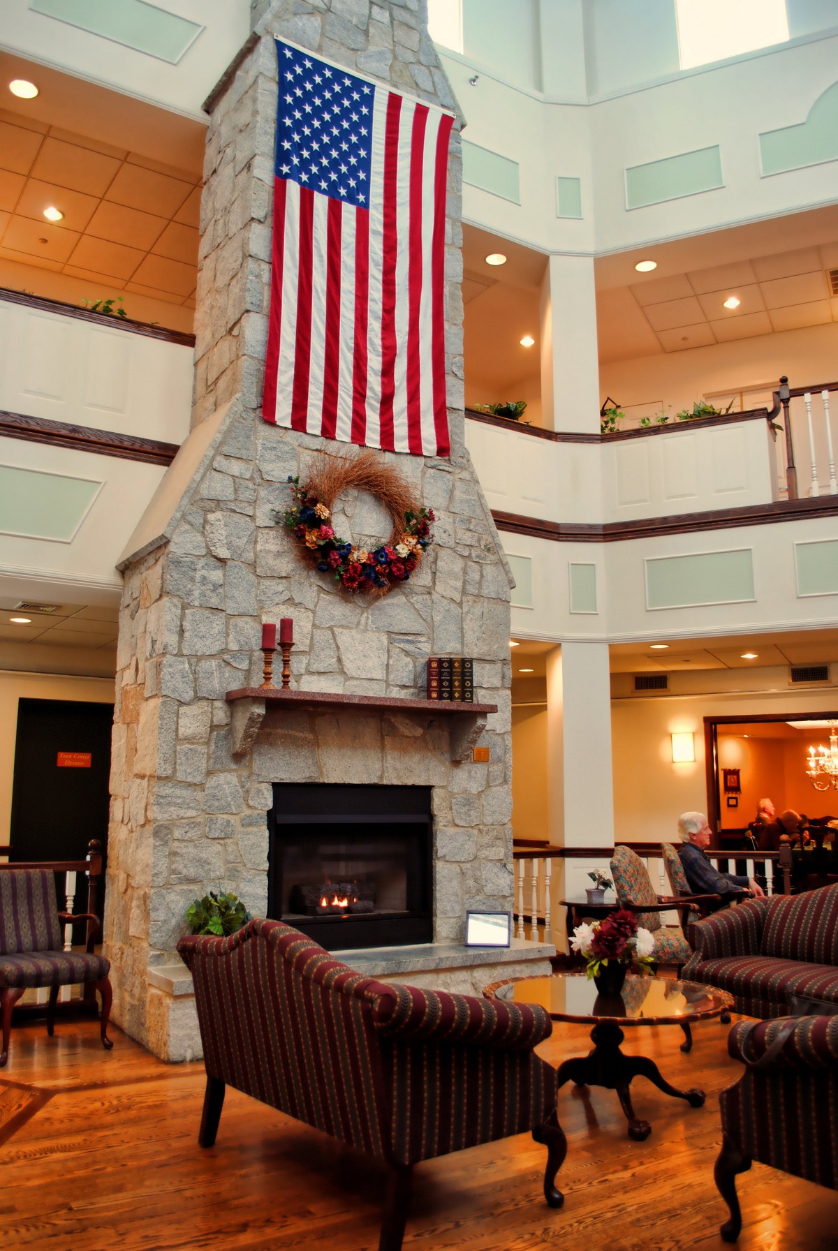 A cozy lobby with a central stone fireplace, adorned with an American flag and a decorative wreath, surrounded by comfortable seating.