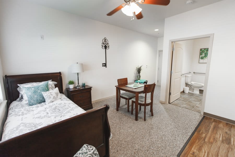 A cozy bedroom with a wooden bed, a small dining table with chairs, and a ceiling fan overhead.