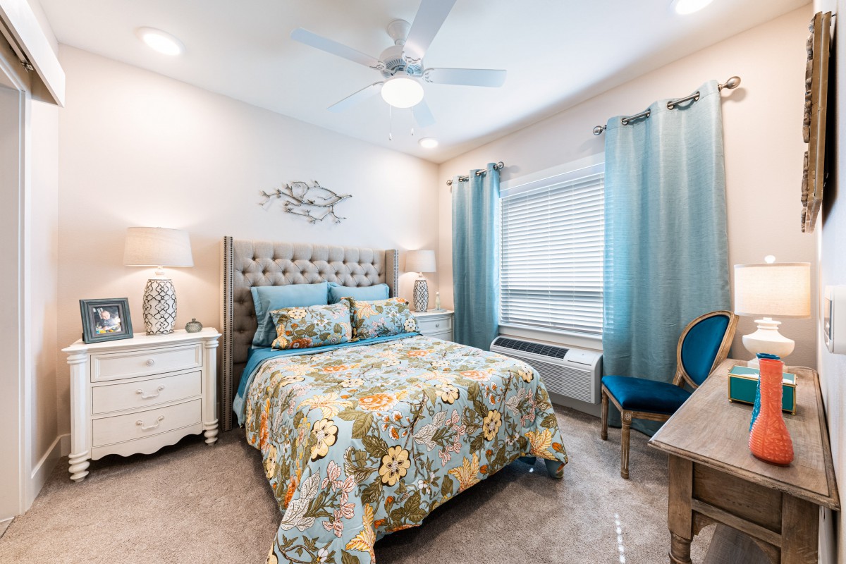 A cozy bedroom with floral bedding and blue accents, featuring a ceiling fan and natural light through the window.
