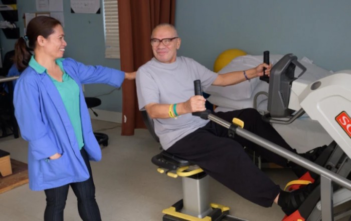 An older adult using a rowing machine with assistance in a gym setting.