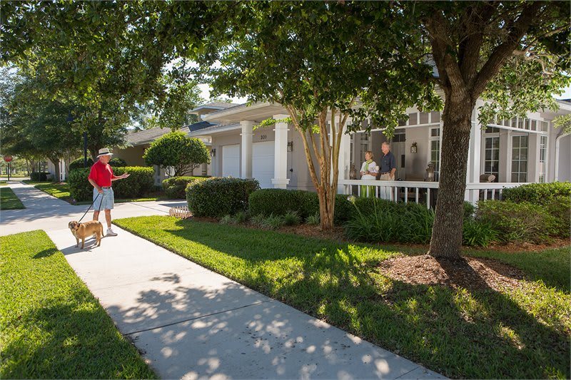A suburban house with a well-maintained lawn and people walking on the sidewalk under the sunny sky.