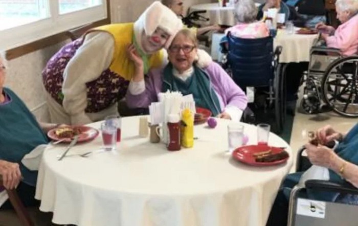 A cheerful clown entertains a senior at a nursing home dining area.