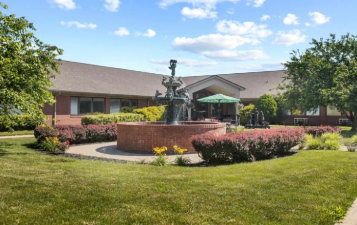 A scenic view of a building with a central fountain surrounded by lush landscaping under a partly cloudy sky.