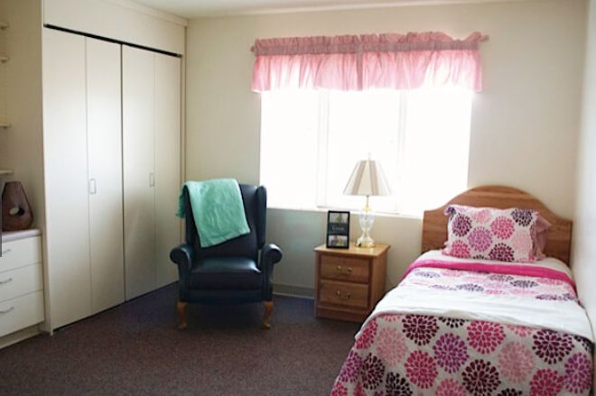 A cozy bedroom featuring a bed with colorful bedding, a nightstand with a lamp, and an armchair by the window.