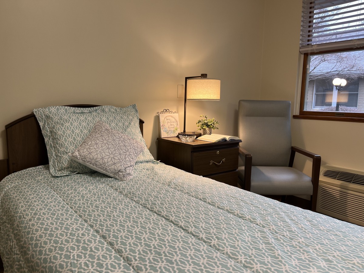 A cozy bedroom with a neatly made bed, a lamp on the nightstand, and an armchair by the window.