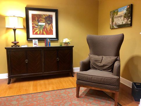 A cozy interior room with a high-back chair, a wooden cabinet, framed artwork, and a lamp.