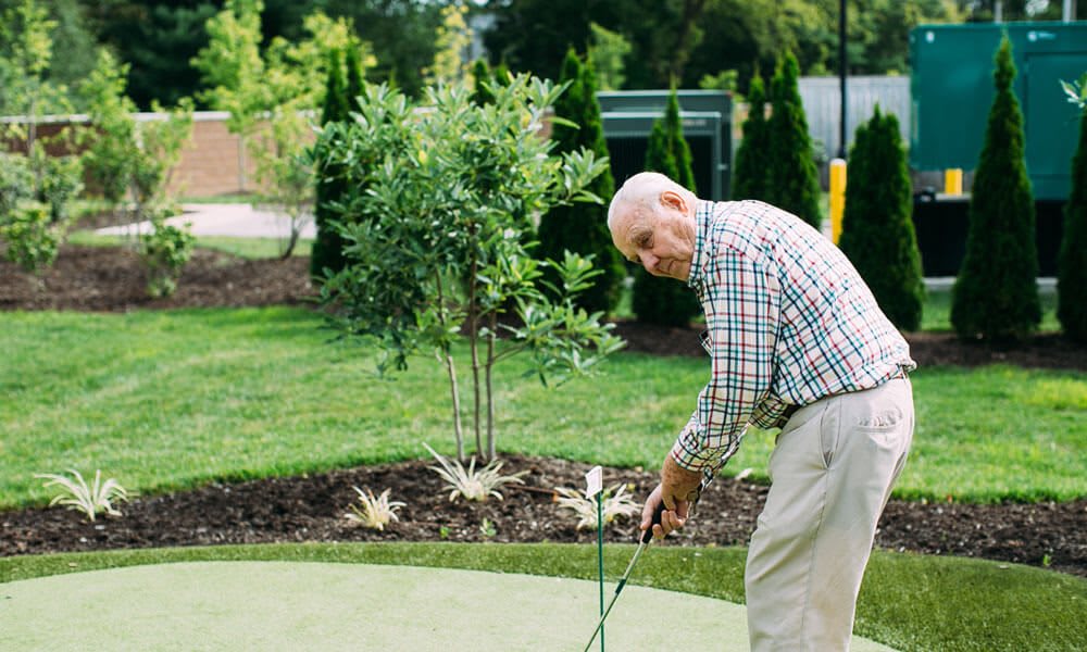 An elderly man practicing his golf putt on a sunny day outdoors.