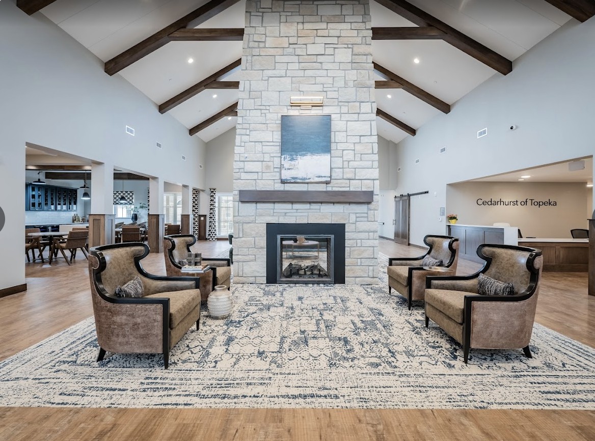 A spacious and cozy living room with a central stone fireplace, surrounded by elegant furniture and high vaulted ceilings with exposed beams.