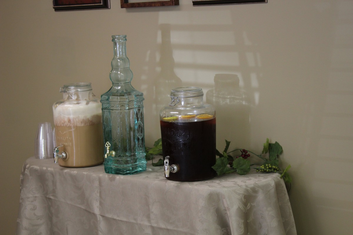 A table with glass beverage dispensers containing various drinks, set up indoors.