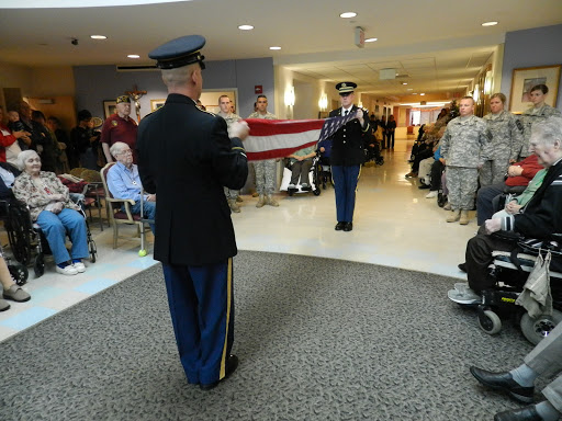 A military honor guard presents the flag in a ceremonial setting, attended by members of the community.