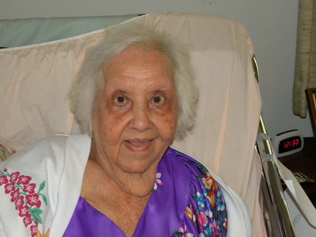 An elderly woman smiling while seated indoors, wearing a purple outfit.