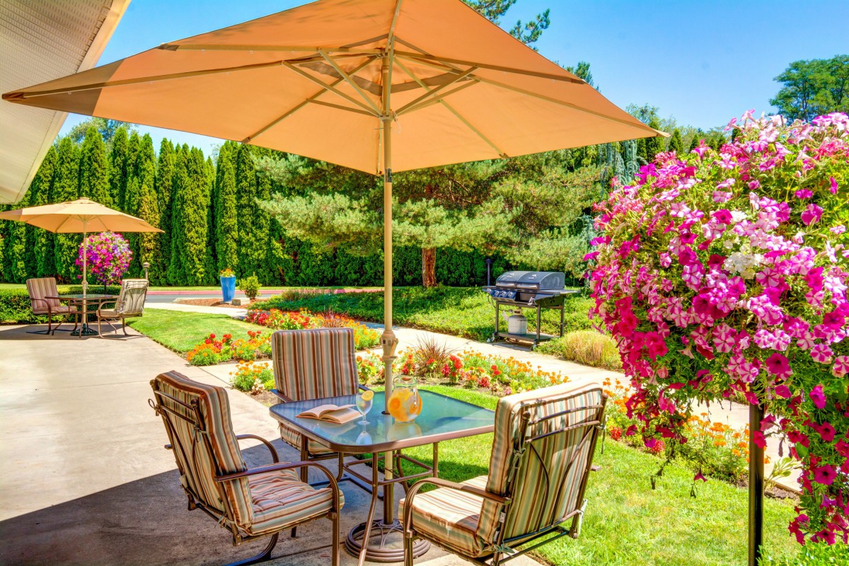 A picturesque garden patio with a table and chairs under a large umbrella, surrounded by lush greenery and vibrant flowers.
