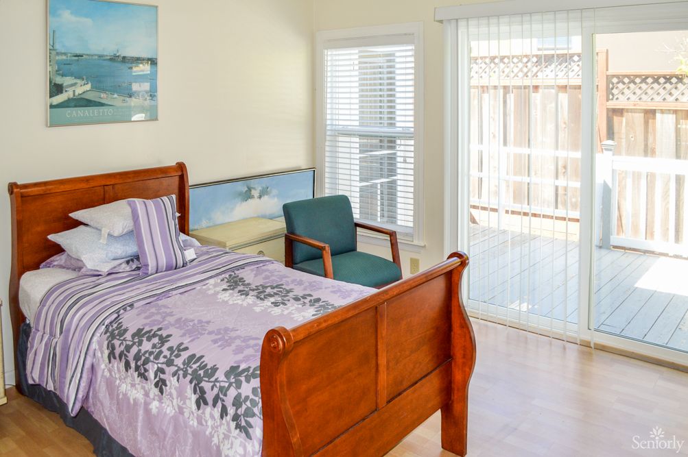 A cozy bedroom featuring a wooden bed, chair, and large windows with blinds.