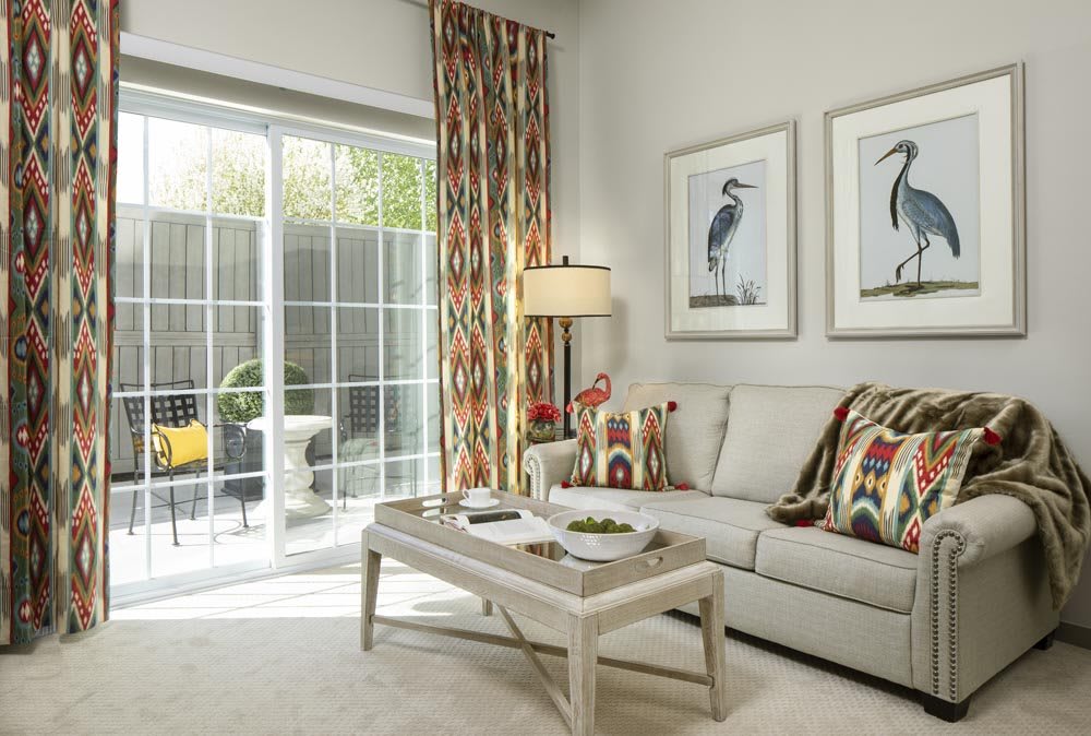 A cozy living room with a neutral sofa, colorful pillows, and artwork featuring birds on the walls. Natural light streams in through a sliding glass door, illuminating the space.