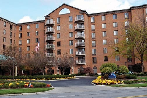 A brick apartment building with balconies, surrounded by landscaped gardens at the entrance.
