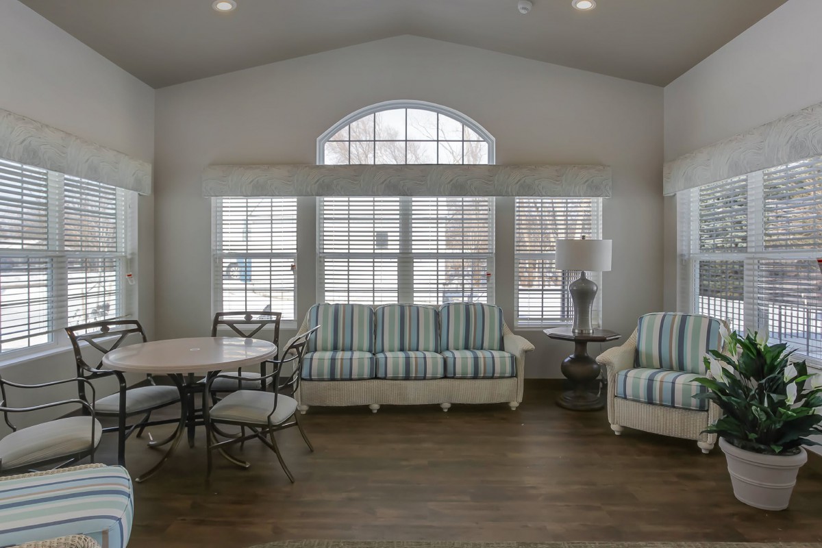 A well-lit living room with large windows, featuring striped sofas and a round table with chairs on hardwood floors.