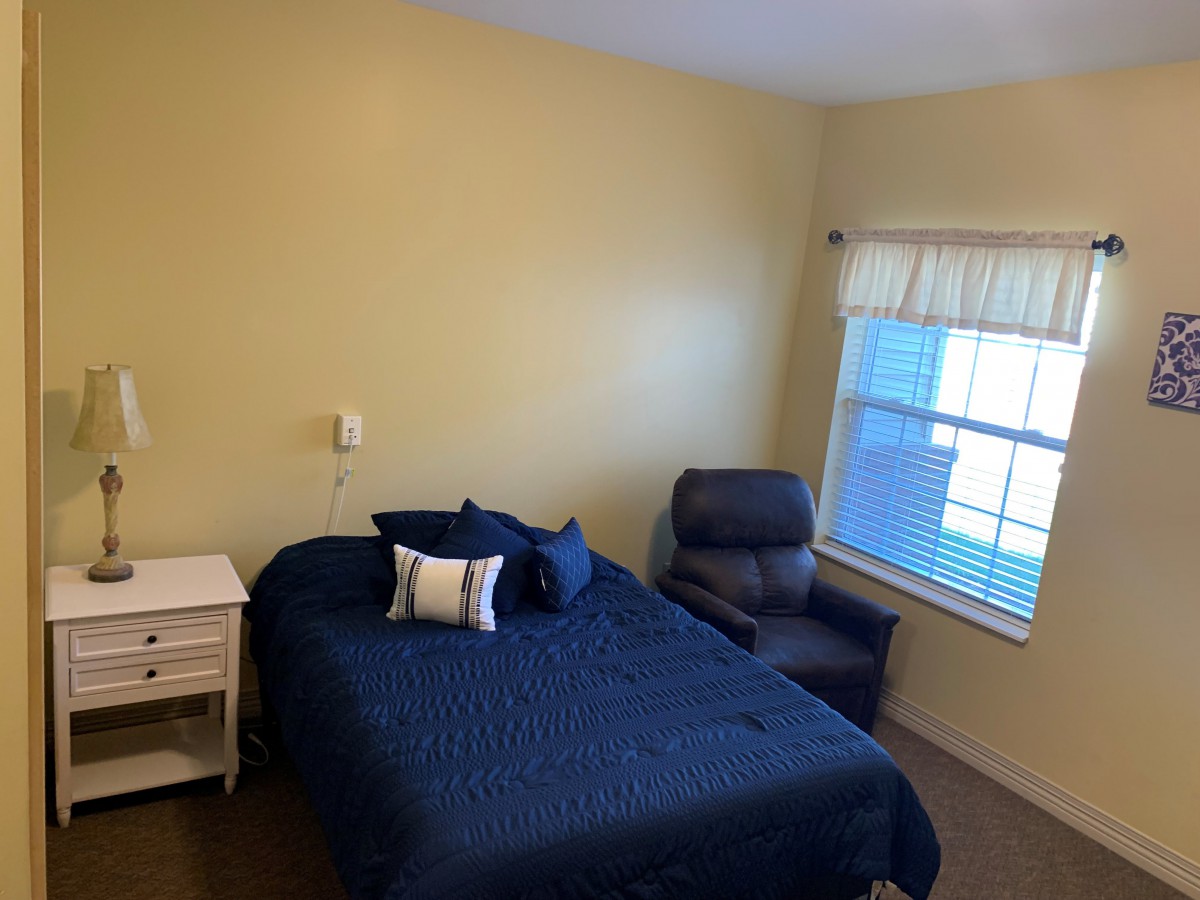 A cozy bedroom with a bed covered in blue bedding, an armchair by the window, and a nightstand beside the bed.