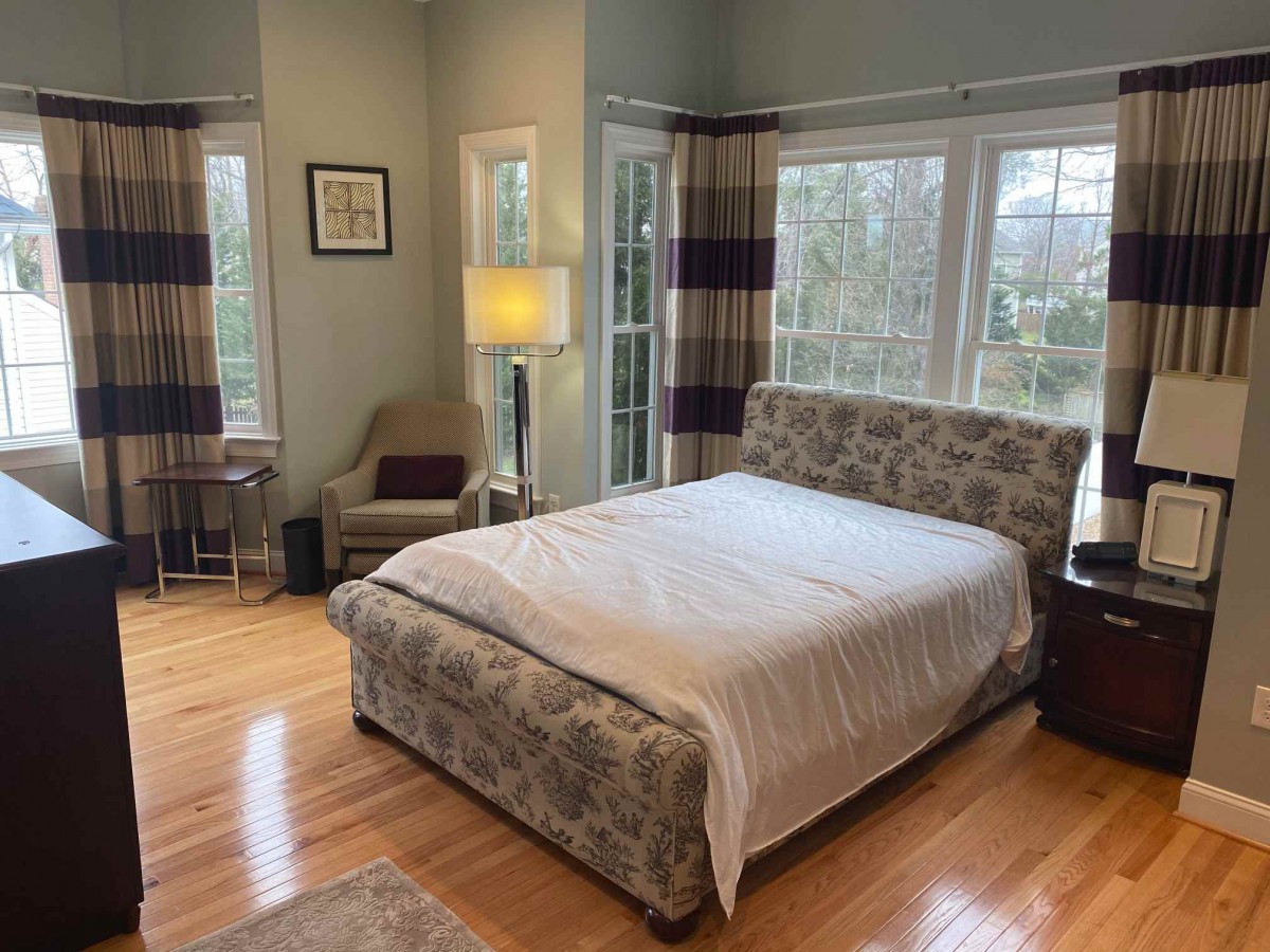 A cozy bedroom with a large bed, surrounded by windows and complemented by a comfortable chair and a standing lamp.