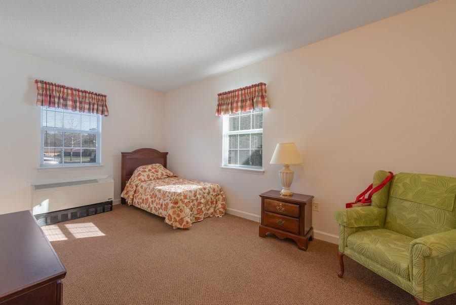A bright bedroom with a single bed, nightstand, lamp, and an armchair next to two windows.