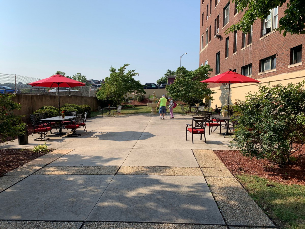 A sunny outdoor patio with tables, chairs, and red umbrellas next to a building.