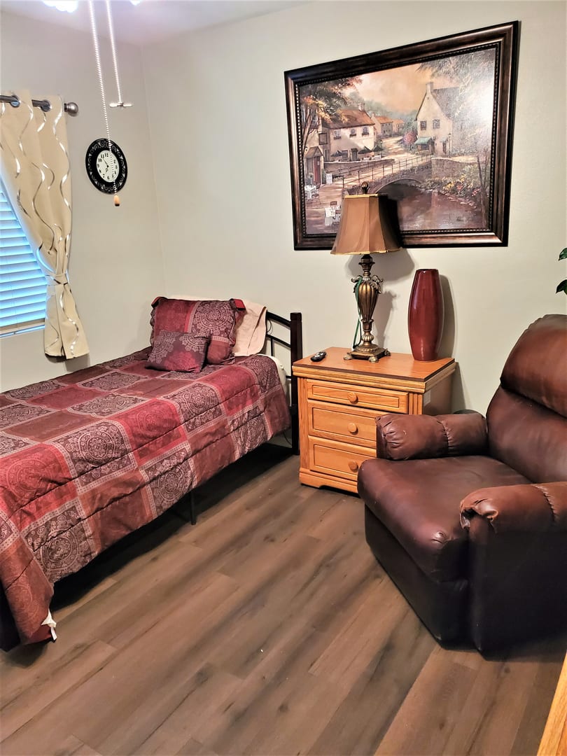 A cozy bedroom scene featuring a single bed with red plaid bedding, a dark brown armchair, and a wooden nightstand topped with a lamp. A landscape painting hangs above the nightstand, and a clock adorns the wall beside the window.
