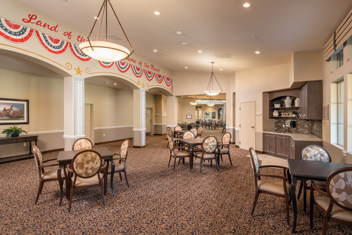 A spacious dining area with elegant chandeliers, decorated ceiling trim, and multiple tables and chairs in a neutral-toned setting.