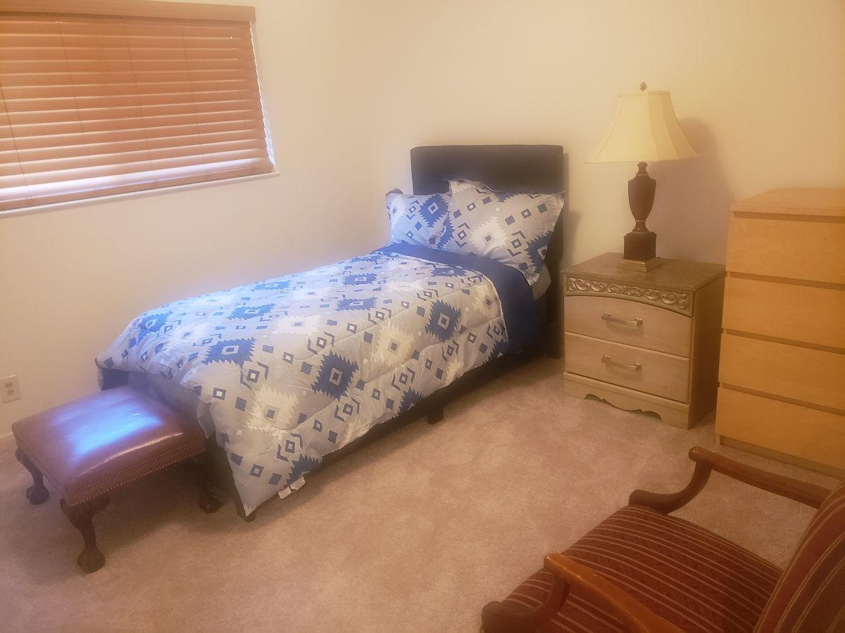 A cozy bedroom featuring a single bed with floral bedding, a wooden nightstand with a lamp, and carpeted flooring.