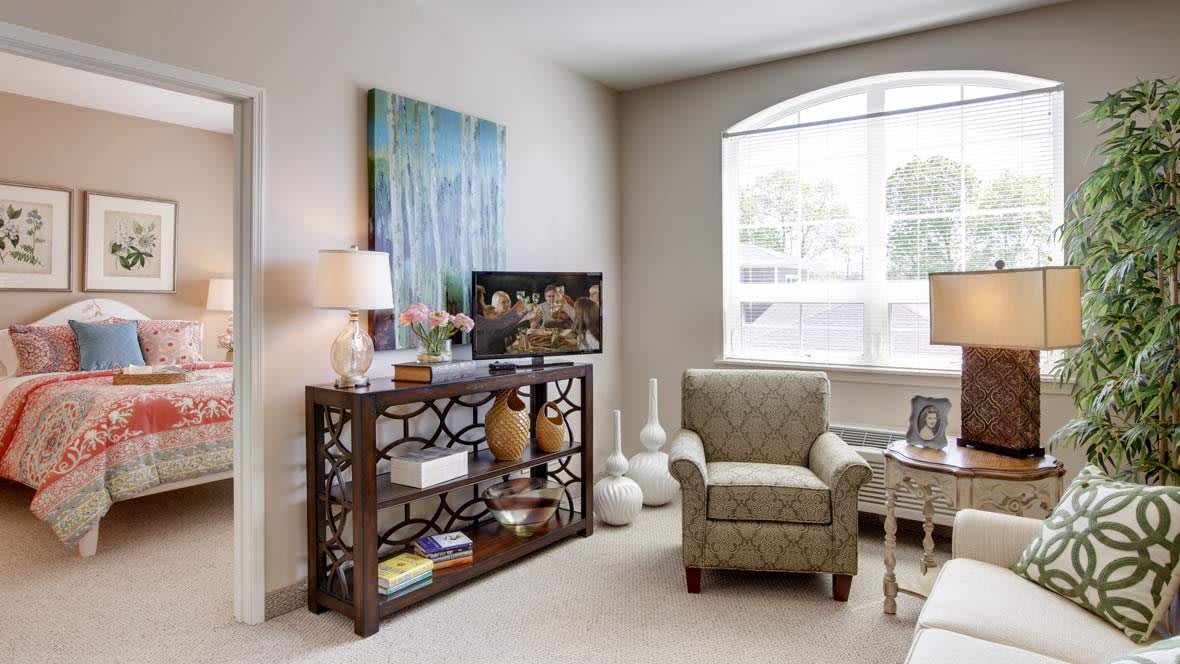 A cozy living room featuring a stylish armchair, decorative bookshelf with vases, and a large window allowing natural light.