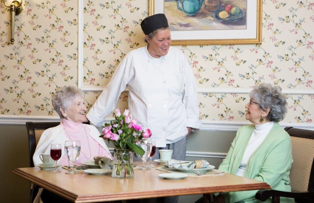 A chef engages in conversation with two elderly women seated at a dining table.