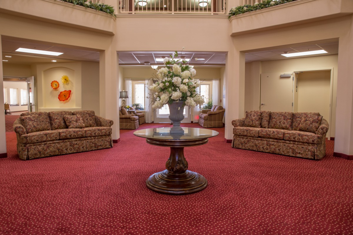 A spacious lobby area featuring a central table with a floral arrangement, surrounded by comfortable sofas and carpeted flooring.