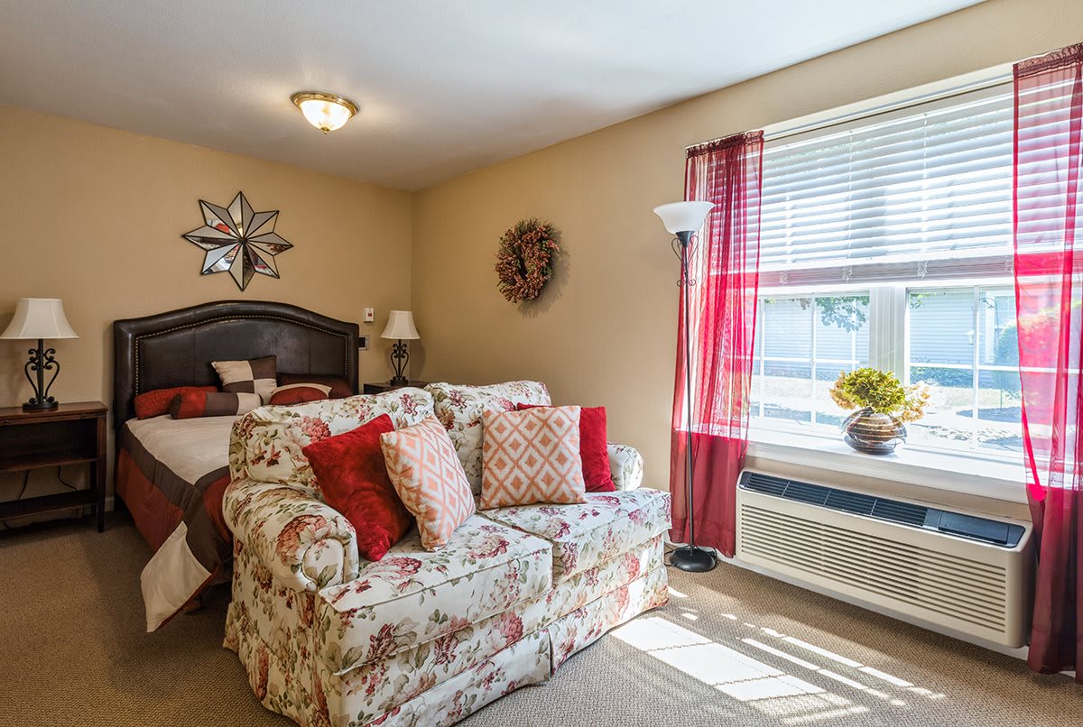 A cozy bedroom featuring a floral-patterned sofa, large bed, and bright natural light streaming through the window with red curtains.