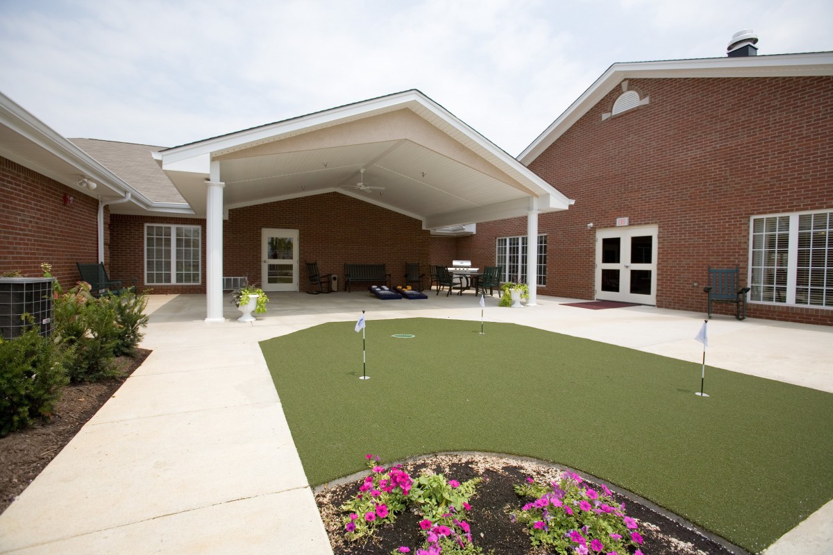 A covered patio area with a small putting green and landscaped garden in front of a brick building.